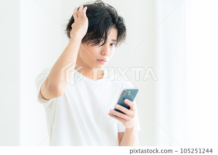 一位年輕人在客廳窗邊一邊操作智能手機一邊護理頭髮 105251244