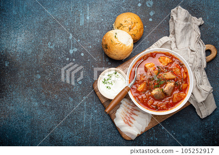 Ukrainian Borscht, red beetroot soup with meat, in white bowl with sour cream, garlic buns Pampushka and salo slices, rustic stone background. Traditional authentic dish of Ukraine, space for text 105252917