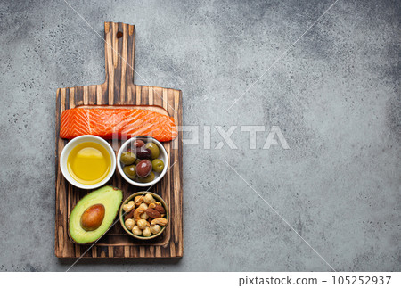 Food sources of healthy unsaturated fat and omega 3: fresh raw salmon fillet, avocado, olives, nuts on cutting board, rustic stone background top view. Healthy nutrition, keto diet, space for text 105252937