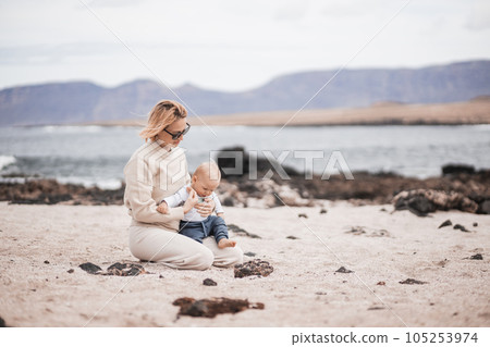 Mother enjoying winter beach vacations playing with his infant baby boy son on wild volcanic sandy beach on Lanzarote island, Canary Islands, Spain. Family travel and vacations concept. Mother enjoying winter beach vacations playing with his infant baby boy son on wild volcanic sandy beach on Lanzarote island, Canary Islands, Spain. Family travel and vacations concept. 105253974