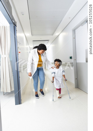 African american female doctor and girl patient walking with crutches in corridor at hospital 105255026