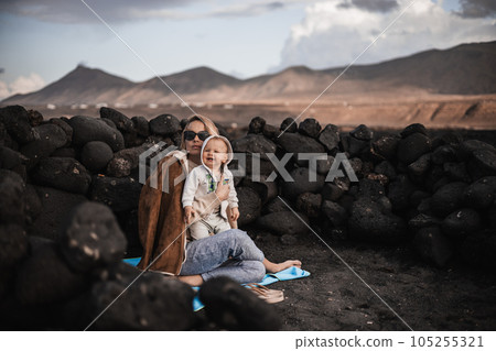 Mother enjoying winter vacations playing with his infant baby boy son on black sandy volcanic beach of Janubio on Lanzarote island, Spain on windy overcast day. Family travel vacations concept 105255321