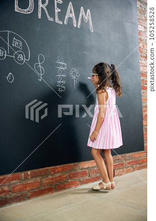 Little girl, child standing at chalkboard and drawing her dreamings, flowers. Imagination. Psychology and creative thinking 105258528