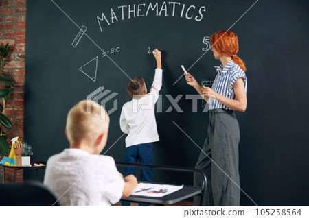 Little bot, child, student standing at chalkboard, writing and solving math tasks with teacher. Lessons at school 105258564