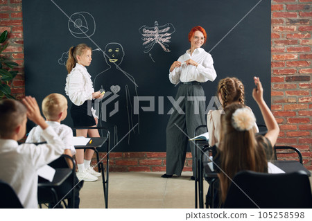 Teacher of primary school teaching to kids anatomy in classroom. Drawing human shape on blackboard. Kids raising hands to answer 105258598