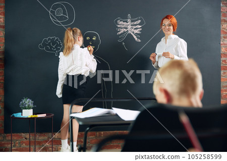 Little girl, child, student of primary school learning anatomy with teacher in classroom. Drawing human shape on blackboard Little girl, child, student of primary school learning anatomy with teacher in classroom. Drawing human shape on blackboard 105258599