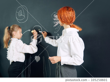 Girl, student of primary school standing at chalkboard with teacher and drawing human representation. Anatomy lessons Girl, student of primary school standing at chalkboard with teacher and drawing human representation. Anatomy lessons 105258601