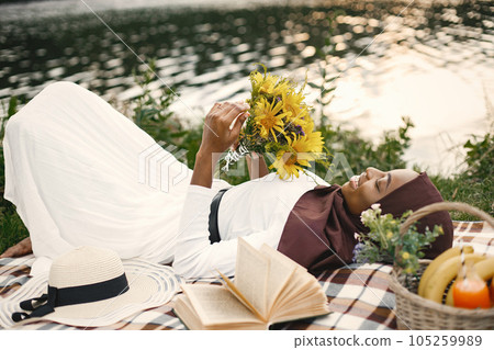 Muslim girl in hijab lay holding a bouquet of flowers. Portrait of a Muslim lady in a brown hijab on the plaid picnic blanket near the river. Girl wearing white clothes. 105259989