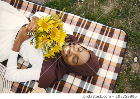 Muslim girl in hijab lay holding a bouquet of flowers. Portrait of a Muslim lady in a brown hijab on the plaid picnic blanket near the river. Girl wearing white clothes. Muslim girl in hijab lay holding a bouquet of flowers. Portrait of a Muslim lady in a brown hijab on the plaid picnic blanket near the river. Girl wearing white clothes. 105259990