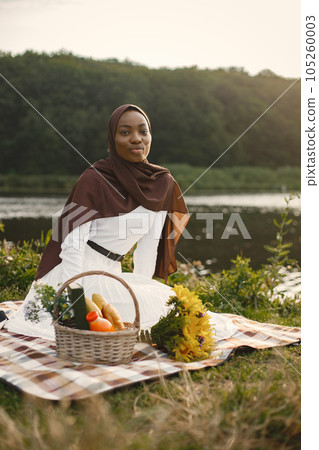 Muslim girl in hijab sits next to a basket with a picnic food and a bouquet of flowers. Portrait of a Muslim lady in a brown hijab on the plaid picnic blanket near the river. Girl wearing white Muslim girl in hijab sits next to a basket with a picnic food and a bouquet of flowers. Portrait of a Muslim lady in a brown hijab on the plaid picnic blanket near the river. Girl wearing white 105260003