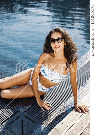 Young girl in a sunglasses sitting near swimming pool. Brunette girl wearing blue and white swimwear. Girl with curly hair posing for a photo near the pool. 105260176