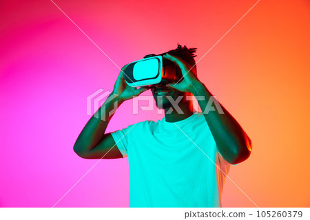 Half length image of african man smiling while playing video games in VR-glasses over pink-orange background in neon. 105260379