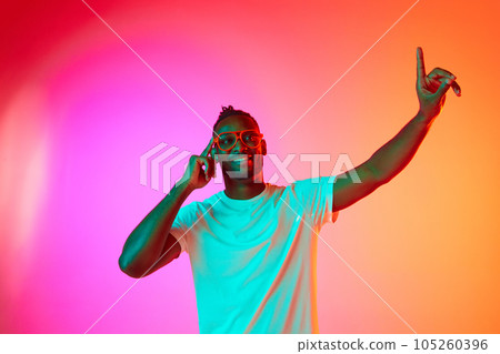 Portrait of smiling african man looking at camera and moves his hands to the side over gradient colorful background in neon. Portrait of smiling african man looking at camera and moves his hands to the side over gradient colorful background in neon. 105260396