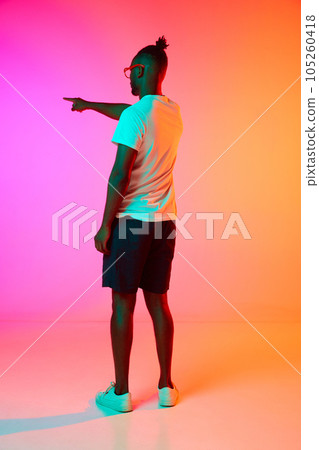 Full length image of backview african man in glasses holding his hand up and pointing something over orange-pink background in neon. Full length image of backview african man in glasses holding his hand up and pointing something over orange-pink background in neon. 105260418