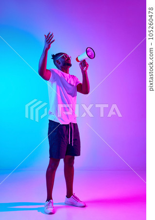 Full length questioning african man shouting in megaphone, looking up and holding hands up over colorful studio background in neon. 105260478