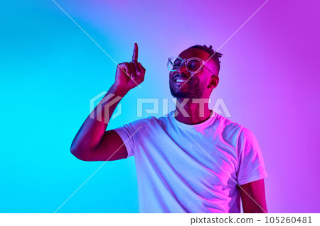 Studio shot of african-american smiling man in glasses looking away holding his finger up over gradient colorful background in neon. Studio shot of african-american smiling man in glasses looking away holding his finger up over gradient colorful background in neon. 105260481