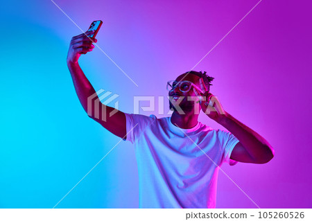 Photo with african-american man with dreadlocks taking selfie on phone and showing v-sign over gradient blue-pink background in neon 105260526