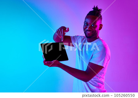 Young african man with afro hairdo holding padlet pointing at gadget screen and looking at camera over gradient blue-pink background in neon. 105260550