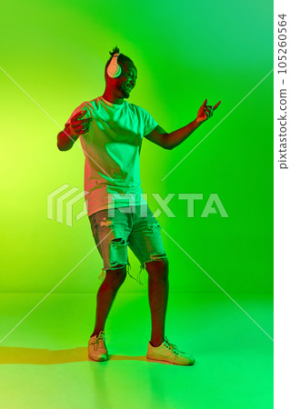 Full length image of dancing young man with dreadlocks in headphones with raising hand up over gradient colorful background in neon. 105260564