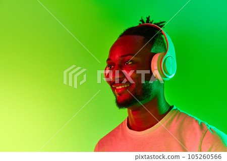 Image of young african man looking away in headphones and smiling over gradient colorful background in neon. 105260566