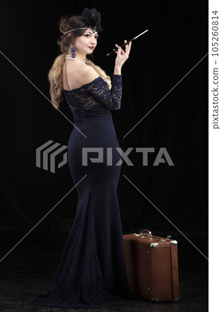 A beautiful girl dressed in the style of the 20-30s of the last century with a cigar and a suitcase on a dark background. Vintage retro girl. 105260814