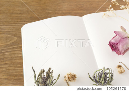 A white notebook on a wooden desk with a decorative frame composed of dried flowers such as pink roses on it 105261578