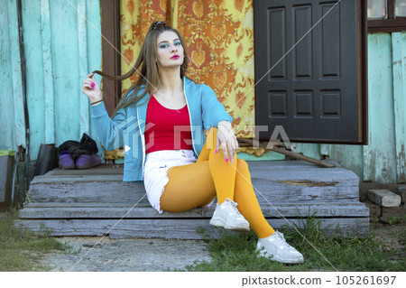 A beautiful girl in bright clothes sits on the porch of a village house. Woman in the style of the 90s. A beautiful girl in bright clothes sits on the porch of a village house. Woman in the style of the 90s. 105261697