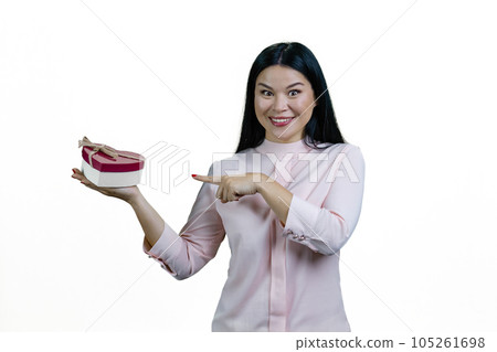 Young asian brunette woman is pointing at heart-shaped gift box on her palm. Valentines day or anniversary. Isolated on white background. Young asian brunette woman is pointing at heart-shaped gift box on her palm. Valentines day or anniversary. Isolated on white background. 105261698