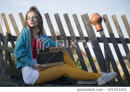 A beautiful country girl in bright clothes sits on a wooden bench with a cassette recorder. Woman in the style of the 90s. A beautiful country girl in bright clothes sits on a wooden bench with a cassette recorder. Woman in the style of the 90s. 105261842
