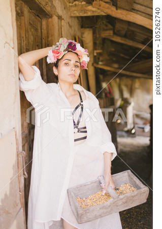 Beautiful indian woman in a white dress and a wreath on her head holds a basket with corn. The farm or countryside Beautiful indian woman in a white dress and a wreath on her head holds a basket with corn. The farm or countryside 105262740