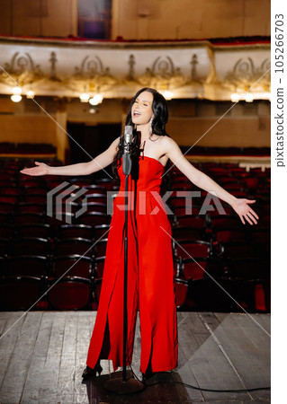 A luxurious actress in a red dress sings on stage with a microphone in her hands A luxurious actress in a red dress sings on stage with a microphone in her hands 105266703