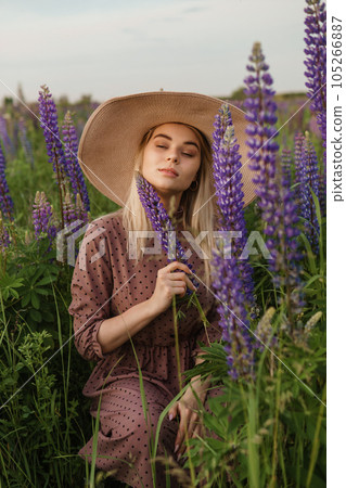 A beautiful woman in a straw hat walks in a field with purple flowers. A walk in nature in the lupin field A beautiful woman in a straw hat walks in a field with purple flowers. A walk in nature in the lupin field 105266887