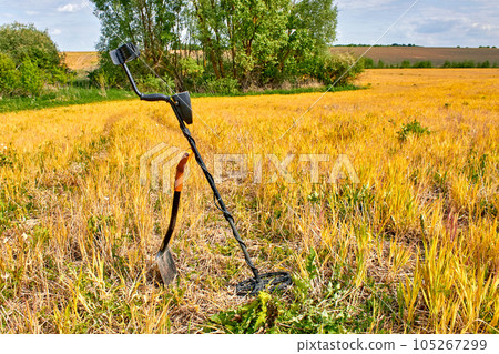 Metal detector and shovel for searching for coins and precious metals against background of yellow field and green forest Metal detector and shovel for searching for coins and precious metals against background of yellow field and green forest 105267299