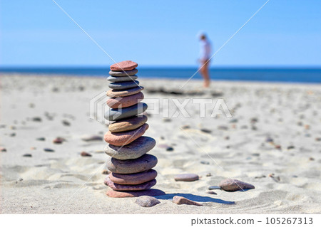Balanced pyramid of stones on sandy beach against backdrop of bustle of woman collecting stones on seashore Balanced pyramid of stones on sandy beach against backdrop of bustle of woman collecting stones on seashore 105267313
