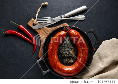 Fried pork sausage tied with a ring on a black cast iron pan on a wooden cutting board with hot peppers and cutlery on a black stone table. Top view with copy space Fried pork sausage tied with a ring on a black cast iron pan on a wooden cutting board with hot peppers and cutlery on a black stone table. Top view with copy space 105267315