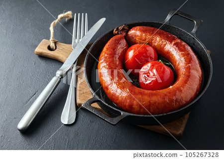 Grilled beef sausage tied with a ring and baked tomatoes on a black cast iron pan on a wooden cutting board with cutlery on a black stone table Grilled beef sausage tied with a ring and baked tomatoes on a black cast iron pan on a wooden cutting board with cutlery on a black stone table 105267328
