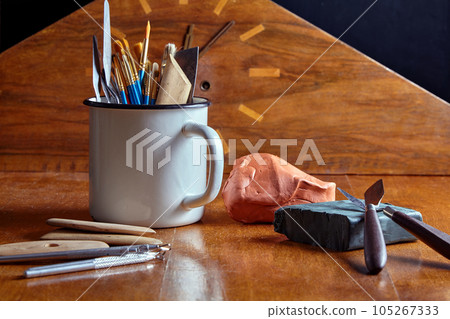 Tools for modeling in white metal mug and pieces of green and red plasticine and clay against background of mantel clock Tools for modeling in white metal mug and pieces of green and red plasticine and clay against background of mantel clock 105267333