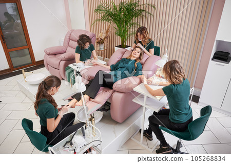 Woman relaxing in comfortable salon chair while having manicure, pedicure and eyelash extension procedure. Manicurist, pedicurist and eyelash specialist working with client in modern beauty salon. Woman relaxing in comfortable salon chair while having manicure, pedicure and eyelash extension procedure. Manicurist, pedicurist and eyelash specialist working with client in modern beauty salon. 105268834
