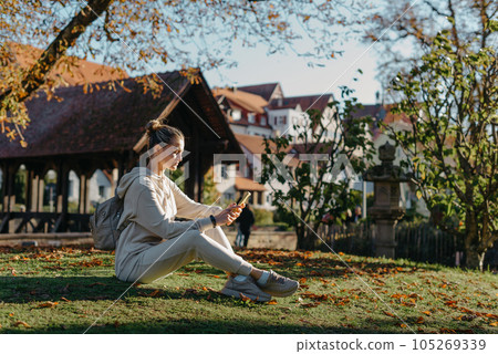 Young Fashionable Teenage Girl With Smartphone In Europian Park In Autumn Sitting At Smiling. Trendy Young Woman In Fall In Park Texting. Retouched, Vibrant Colors. Beautiful Blonde Teenage Girl 105269339