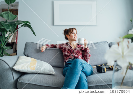 Happy smiling proud of herself young woman relaxing with coffee cup after she puts by her own a picture on the wall at her home. Housekeeping work. Doing repair herself. DIY, gender equality concept. 105270904