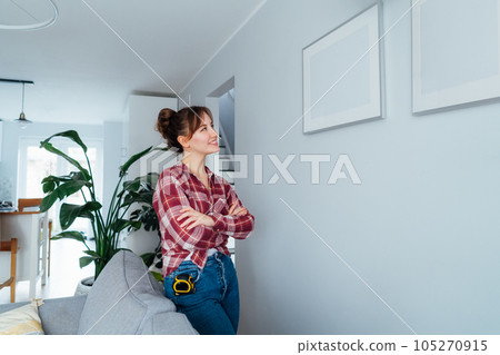 Happy smiling proud of herself young brunette woman after she put by her own picture mockups on the wall at her home. Housekeeping work. Doing repair herself. DIY, gender equality concept Happy smiling proud of herself young brunette woman after she put by her own picture mockups on the wall at her home. Housekeeping work. Doing repair herself. DIY, gender equality concept 105270915