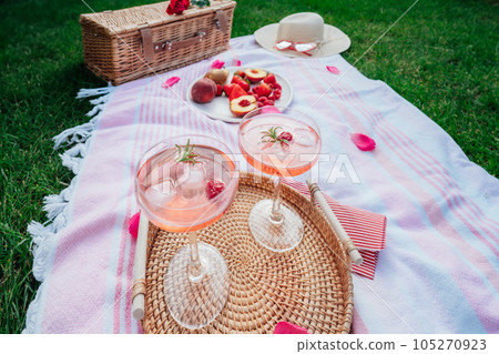 Pink drinks, cocktail with ice, raspberry, rosemary. Two glasses with martini, champagne, cider, lemonade on the blanket with fruit plate, picnic basket. Cozy summer picnic on nature. Selective focus 105270923