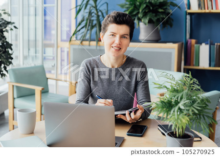 Smiling neutral gender middle-aged woman making notes in her paper notebook, using laptop while working indoors in her workstation in an open space office. Online video call, IT HR, recruiting 105270925