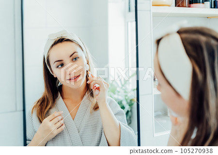 Young woman in bathrobe looking in mirror and making face massage with jade roller in the bathroom. The concept of natural procedures for skin care. Home Beauty self-care treatment. Selective focus 105270938
