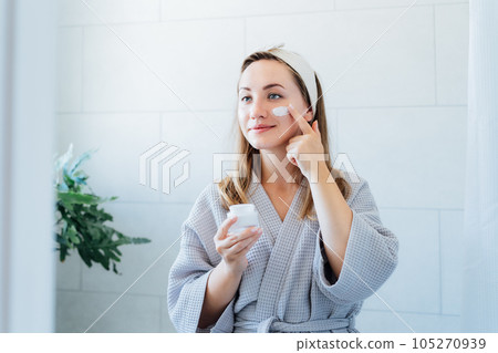 Young woman in bathrobe looking in the mirror, holding bottle with moisturizer cream and applying it on face. Skin hydrating. Cosmetic spa procedures. Beauty self-care at home. Selective focus. Young woman in bathrobe looking in the mirror, holding bottle with moisturizer cream and applying it on face. Skin hydrating. Cosmetic spa procedures. Beauty self-care at home. Selective focus. 105270939