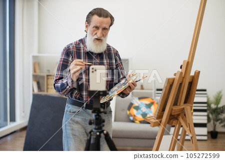 Grey-bearded artist recording video using smartphone indoors Grey-bearded artist recording video using smartphone indoors 105272599