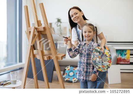 Mom with child's hand and brush standing near easel at home Mom with child's hand and brush standing near easel at home 105272895