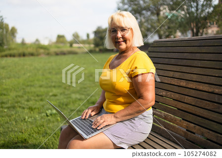 Senior blonde haired business woman using laptop computer, typing on keyboard sitting in park. Stylish freelancer, copywriter working online at workplace. Technology, successful business  Senior blonde haired business woman using laptop computer, typing on keyboard sitting in park. Stylish freelancer, copywriter working online at workplace. Technology, successful business  105274082