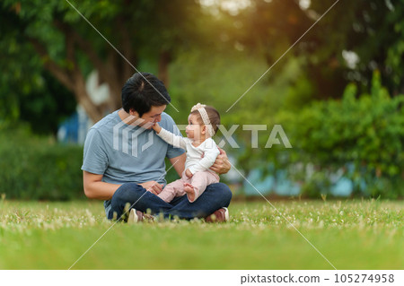 father talking and playing with his infant baby while sitting on grass field 105274958