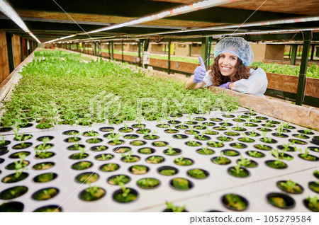 Joyful female gardener showing approval gesture and smiling while leaning on shelf with plant seedling in greenhouse. Woman wearing garden gloves and disposable cap. 105279584